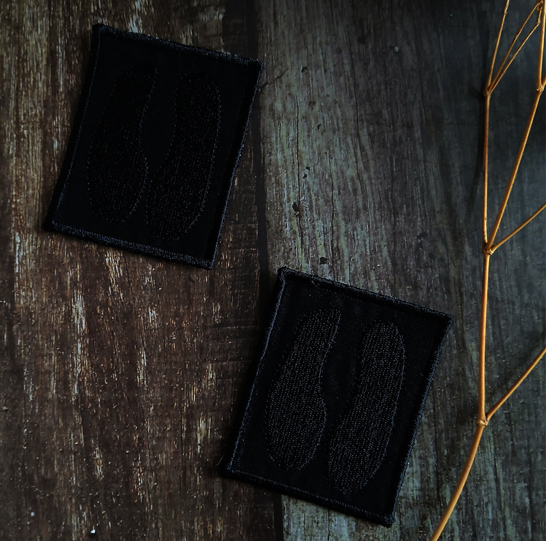 Two black embroidered patches of a cloven hoof on a wooden surface with a dried plant stem.