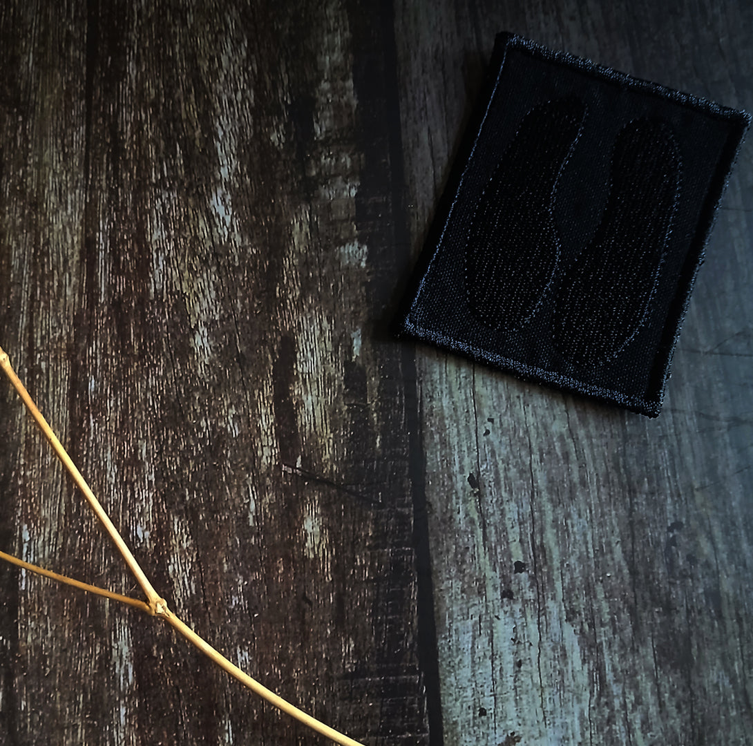 Black embroidered patch on a textured wooden surface with a twig.