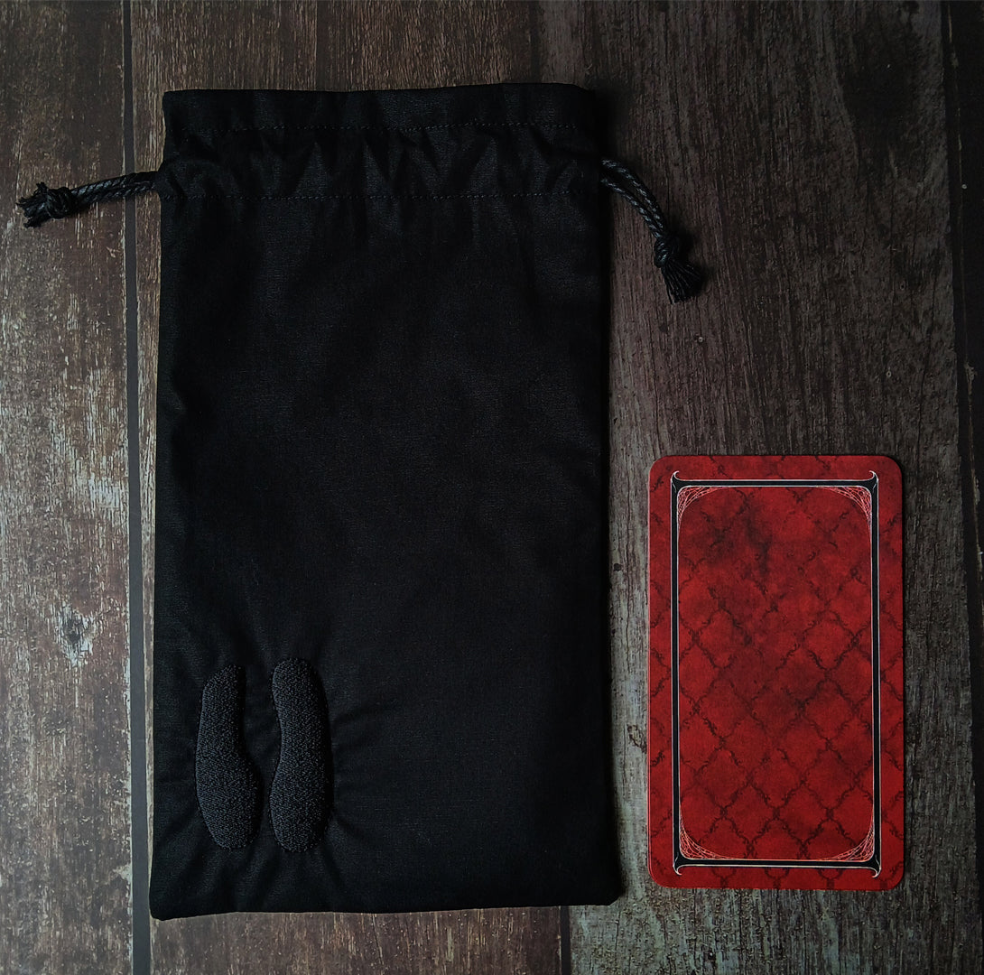 Black drawstring tarot card bag next to red tarot card to show scale on a wooden surface